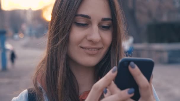 Primer plano retrato de mujer joven las calles de la ciudad soleada, chica alegre usando el teléfono celular, fondo de la puesta del sol . — Vídeos de Stock