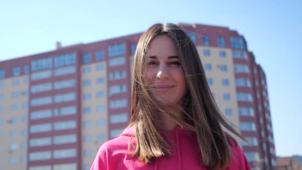 Portret van een donkerharige brunette in een stad op de achtergrond woongebouw. Meisje glimlacht en haar haar ontwikkelt zich in de wind. de zonnen stralen en een warme flits zwerven door het vrouwelijke gezicht. 4k — Stockvideo