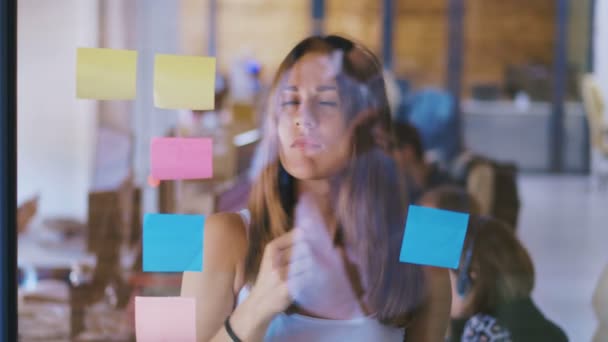 Vista frontal de cerca de la mujer bonita ejecutiva añadiendo notas adhesivas de colores en el vidrio en la oficina moderna — Vídeos de Stock