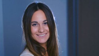 Kahverengi saçlı, büyük kahverengi gözlü, karanlık odada duran mutlu genç kadının yakın görüntüsü, kameraya doğru neşeyle gülümsüyor. Kadın portresi. 4k