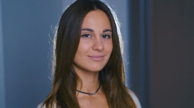 Kahverengi saçlı, büyük kahverengi gözlü, karanlık odada duran mutlu genç kadının yakın görüntüsü, kameraya doğru neşeyle gülümsüyor. Kadın portresi. 4k