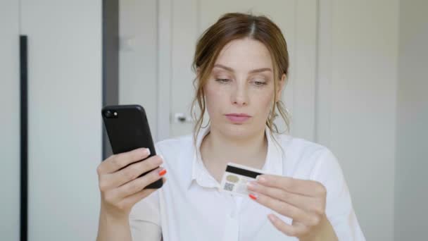 Femme aux cheveux brun clair est assis à la maison et effectue des paiements en ligne via Internet à partir d'une carte de débit sur son smartphone. 4k — Video