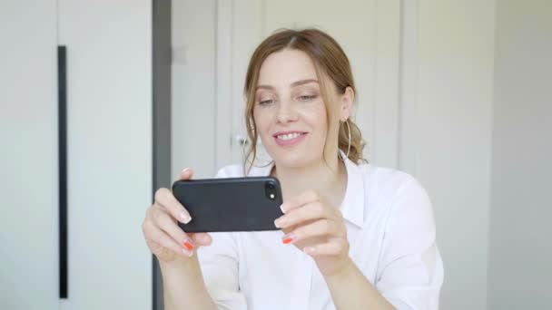 Щаслива розслаблена молода жінка тримає смартфон, дивлячись на екран мобільного телефону, сміючись, користуючись мобільними додатками для покупок, весело граючи в ігри балачки в соціальних мережах сидячи вдома 4k — стокове відео