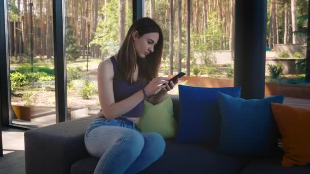 Mujer joven y relajada usando el teléfono inteligente navegando por las redes sociales, revisando noticias, jugando juegos móviles o mensajes de texto sentados en el sofá. — Vídeo de stock