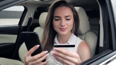Arabada oturan, elinde kredi kartı ve akıllı telefonuyla gülümseyen genç kız müşteri. İnternetten alışveriş yapan mutlu bayan alışverişçi kolay taşınabilir ödemeler yapıyor. E-bankacılık uygulama servisi