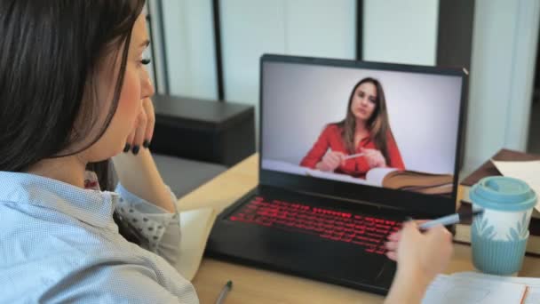 若い学生はオンラインレッスンを見て家から勉強します。教授の後のコンピュータ画面を見ながらメモを取る若い女性は、ビデオ通話のチャートに予測を行います。女子学生 — ストック動画