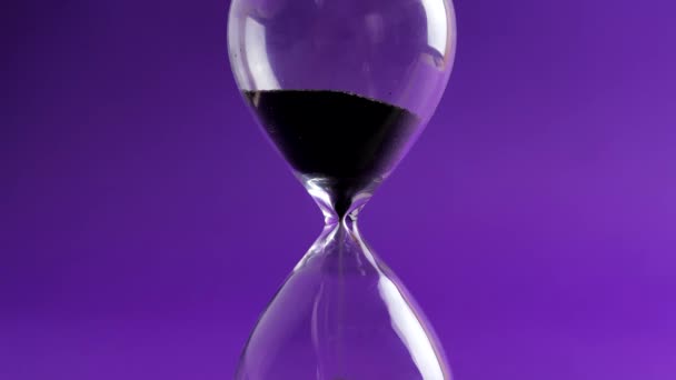 Shot of a sand clock measuring time while the sand is falling down with the purple background - παλιό κλασικό χρονόμετρο. Extreme close up μιας διάφανης κλεψύδρας με ροή μαύρη άμμο - έννοια του χρόνου — Αρχείο Βίντεο