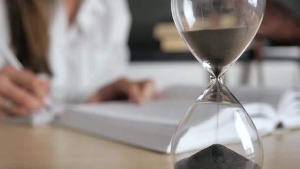 Reloj de arena de primer plano. Silueta mujer leyendo un libro y escribir el prospecto sobre un fondo borroso 4k — Vídeo de stock