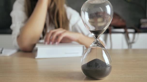 Verter arena en un reloj de arena de pie sobre el escritorio en el espacio de trabajo. Mujer leyendo libros en fondo borroso concepto de educación a distancia 4k — Vídeo de stock