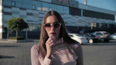 Gözlüklü çekici bir kadın elektronik sigara içiyor, güneşli bir günde, rüzgâr saçlarına esiyor.