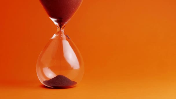 Tournage d'une horloge à sable mesurant le temps pendant que le sable tombe sur le fond orange - vieille minuterie classique. Extrême gros plan d'un sablier transparent avec sable noir coulant - concept de temps — Video