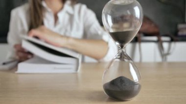 Şeffaf bir mataranın içindeki kum saati, arka planda kitap okuyan bir kızın silüeti dökülüyor ve zamanın geçişini noktalıyor.