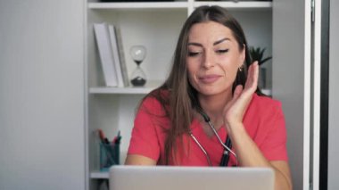 Mercan cüppeli genç beyaz kadın doktor dolapta masada oturuyor ve dizüstü bilgisayarla görüntülü sohbet ediyor. 4k