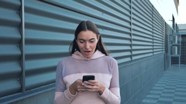Menina atraente nova que olha surpreendente segurando telefone celular conversando e rindo no fundo da cidade. — Vídeo de Stock