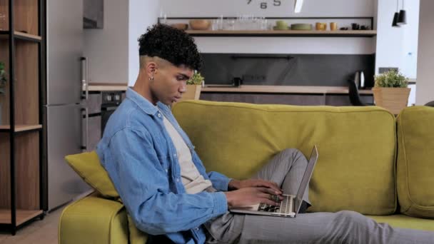 Расслабленный серьезный смешанной расы мужчина подросток фрилансер с помощью ноутбука устройство опираясь на диван в домашнем офисе, целенаправленный предприниматель работает дистанционно печатая на ноутбуке в квартире 4k — стоковое видео