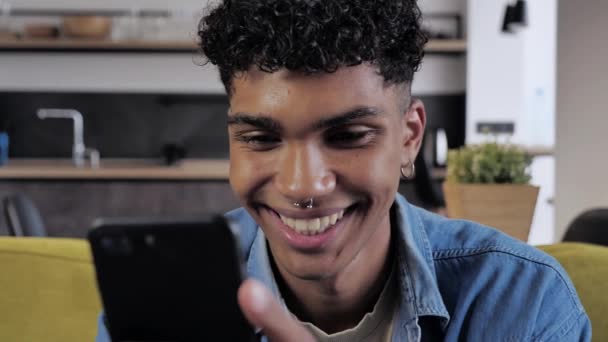 Jovem afro-americano masculino segurando dispositivo smartphone mensagem SMS sms sentado em casa no sofá, sorrindo cara preto usando aplicativos jogando jogo móvel conversando em redes sociais navegar na web no telefone — Vídeo de Stock