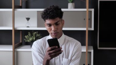 Genç Afro-Amerikan öğrenci elinde akıllı telefon cihazı ile mesaj atıyor modern ofiste oturuyor, sosyal medyada uygulamaları kullanan gülümseyen siyah adam telefonda 4K