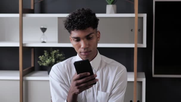Jeune étudiant afro-américain tenant smartphone dispositif SMS message assis au bureau moderne, souriant mec noir en utilisant des applications dans les médias sociaux surf web sur le téléphone à l'intérieur 4k — Video