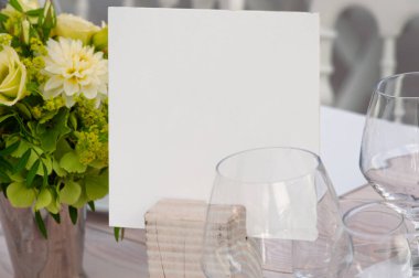 Canlı çiçekler ve şarap kadehleriyle tamamlanmış, ahşap bir tutacağa boş bir kart konan klas bir masa kurulumu. Düğünler, ziyafetler ve zarif etkinlikler için mükemmel..