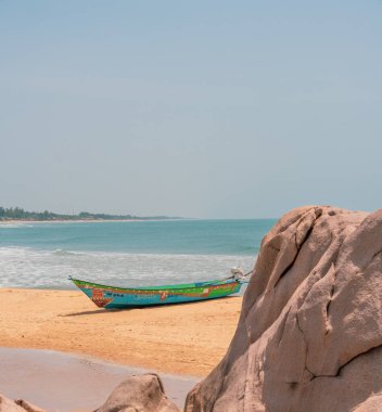 Sahilde büyük bir kayanın yanında küçük bir tekne oturuyor. Gökyüzü açık ve okyanus sakin. Huzurlu ve huzurlu bir sahne.