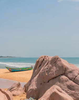 Sahilde büyük bir kayanın yanında bir tekne oturuyor. Gökyüzü açık ve su sakin.