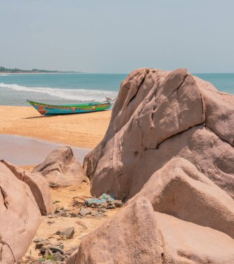 Sahilde büyük bir kayanın yanında bir tekne oturuyor. Kaya, etrafında birkaç küçük kaya bulunan büyük bir kayadır. Kumsal kumlu ve su sakin. Huzurlu ve huzurlu bir sahne.