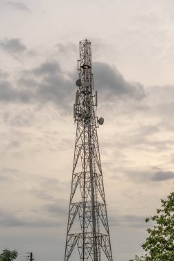 Tepesinde bir sürü anten olan yüksek bir kule. Kule ağaçlarla çevrili. Gökyüzü bulutlu ve güneş batıyor. Tamil Nadu.