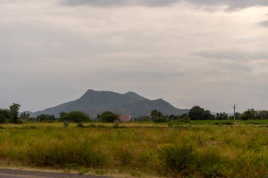 Uzakta bir dağ sırası görülebilir. Gökyüzü bulutlu ve çimenler yeşil. Uzakta bir ev var. Tamil Nadu.