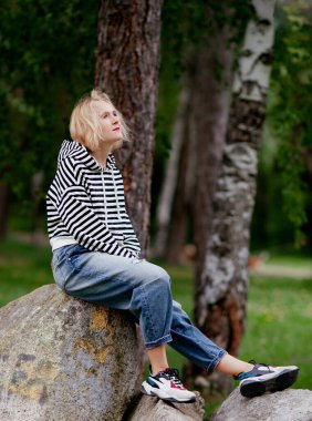 Güzel sarışın kadınlar, sıradan çizgili kapüşonlu ve kot pantolonlu parkta dev bir taşın üzerinde oturuyorlar, meditasyon modunda.