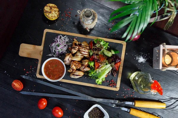 Siyah ahşap arka plan üzerinde bir kesme tahtası üzerinde domates sosu ve yeşillik ile domuz kebap