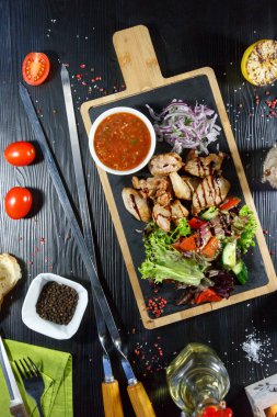 Siyah ahşap arka plan üzerinde bir kesme tahtası üzerinde domates sosu ve yeşillik ile domuz kebap
