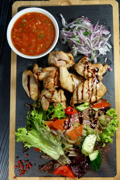 Siyah ahşap arka plan üzerinde bir kesme tahtası üzerinde domates sosu ve yeşillik ile domuz kebap
