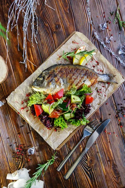 Grilled fish with vegetables and herbs on the kitchen board on a brown wooden background