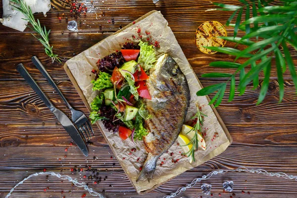 Grilled fish with vegetables and herbs on the kitchen board on a brown wooden background