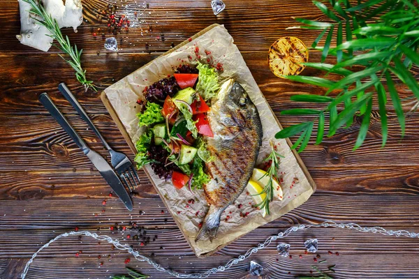 Grilled fish with vegetables and herbs on the kitchen board on a brown wooden background