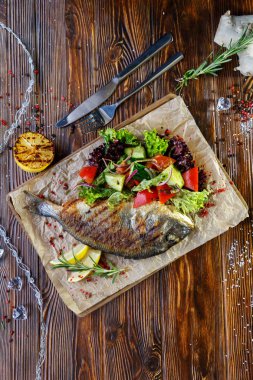 Kahverengi ahşap arka plan üzerinde mutfak tahtası üzerinde sebze ve otlar ile ızgara balık