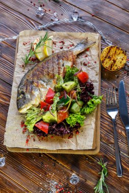 Kahverengi ahşap arka plan üzerinde mutfak tahtası üzerinde sebze ve otlar ile ızgara balık