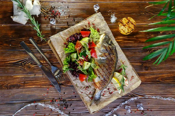 Grilled fish with vegetables and herbs on the kitchen board on a brown wooden background