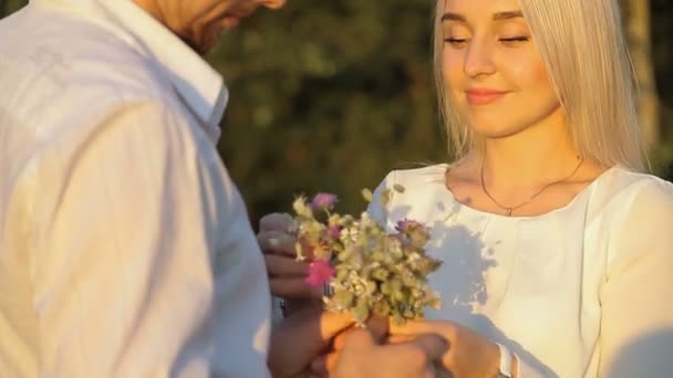 Zijaanzicht van lachende vrouw met bouquet van veldbloemen en vriendje in de buurt van door in veld — Stockvideo