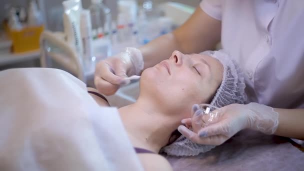 Jovem cliente feminina recebe procedimento facial de beleza. renovação facial. Esteticista usa botão de algodão para cobrir a pele do rosto com máscara de beleza — Vídeo de Stock