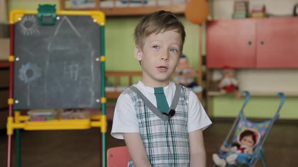 Um menino de 6 anos se senta em uma cadeira com um microfone de lapela em suas roupas e responde a perguntas . — Vídeo de Stock