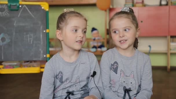 Tweeling meisjes zittend op een stoel beantwoordt vragen. Meisjes geven interviews in een klaslokaal tegen de achtergrond van het Blackboard. — Stockvideo