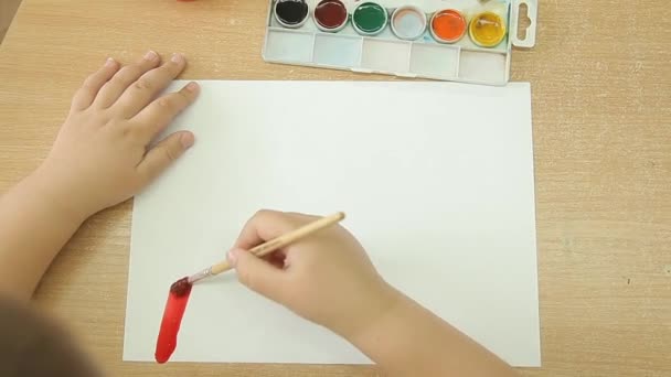 Uma criança segura um pincel na mão direita e desenha um arco-íris em uma folha de papel branca . — Vídeo de Stock