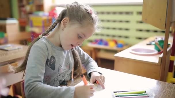 Uma menina desenha um desenho em uma folha branca enquanto se senta em uma mesa no jardim de infância . — Vídeo de Stock