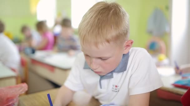 Een roodharige jongen met sproeten tekent een tekening op een wit laken terwijl hij aan een tafel zit in de kleuterschool. — Stockvideo