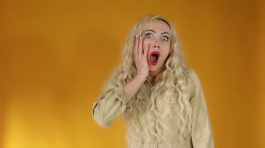 Kafası karışmış Avrupalı sarışın kadın kaşlarını çatmış, elini başının üstünde tutuyor ve kameraya ağzı açık bakıyor, sarı arka planda duran kötü haberi duyduğunda hayal kırıklığına uğruyor..