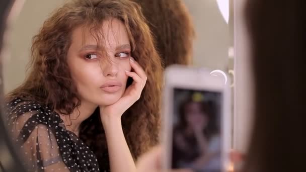 Uma jovem bonita olha para a câmara. Modelo menina posando na frente do telefone da câmera ao tirar uma foto. O estilista do maquiador fotografa o resultado do seu trabalho num telemóvel — Vídeo de Stock