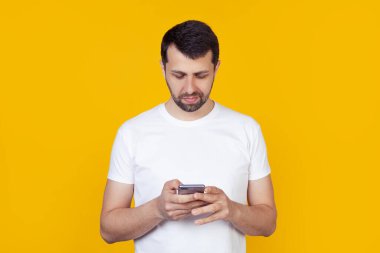 Beyaz tişörtlü, sakallı genç bir adam elinde tuttuğu akıllı telefonuyla internetten bilgi arıyor, doğal bir ifadeyle izole edilmiş sarı bir arka planda duruyor..