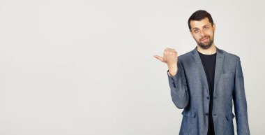 Mutlu bir yüzle gülümseyen sakallı genç bir adam, başparmağıyla reklam için bir yere bakıyor ve işaret ediyor, buraya bakın, izole bir arka planda..