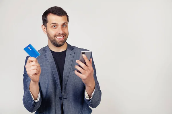 Ceketli sakallı genç bir işadamı, akıllı telefon kullanarak kredi kartını internetten ödeyen yakışıklı bir adam. Gri arka planda bir adamın portresi.
